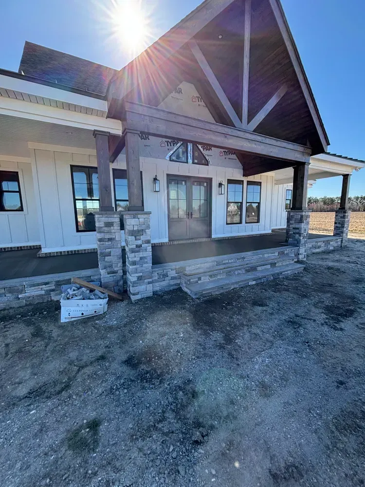 Front porch of a house under construction with stone columns and a sunlit roofline.