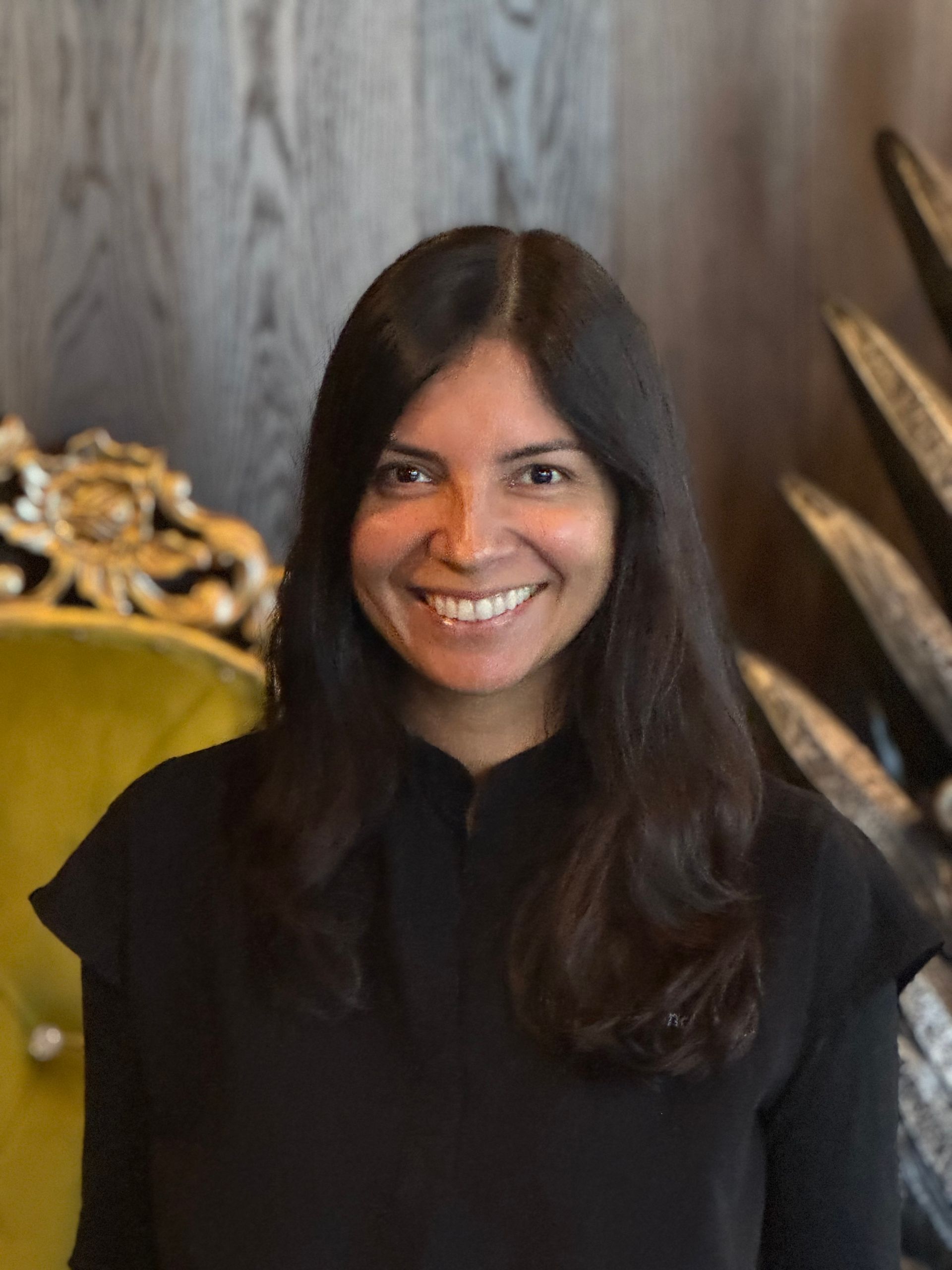 Smiling person with long dark hair in a black top, seated against a wood-paneled wall with a gold chair.