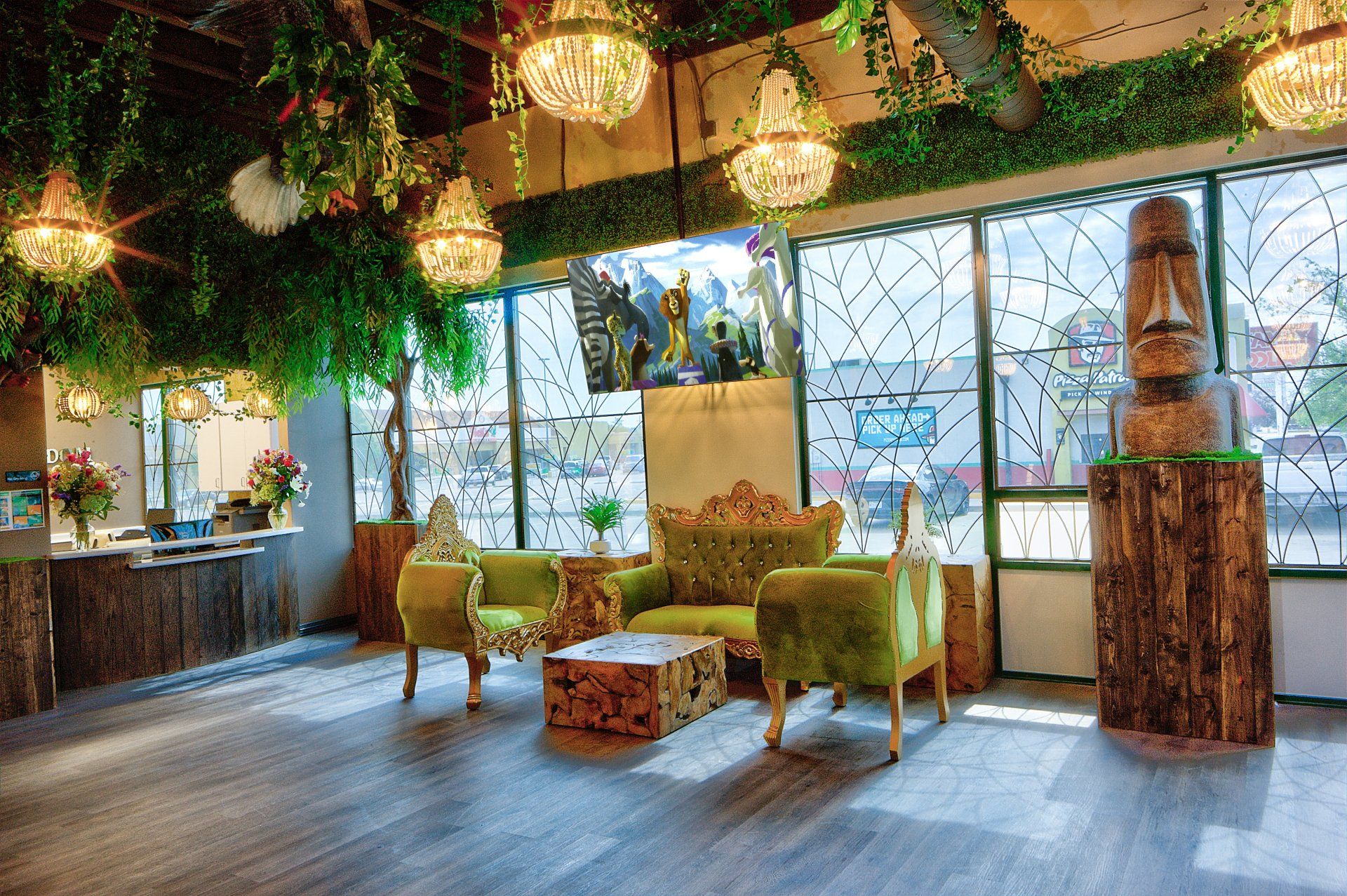 Cozy lounge with green chairs, rustic tables, hanging lights, and leafy decor by large windows