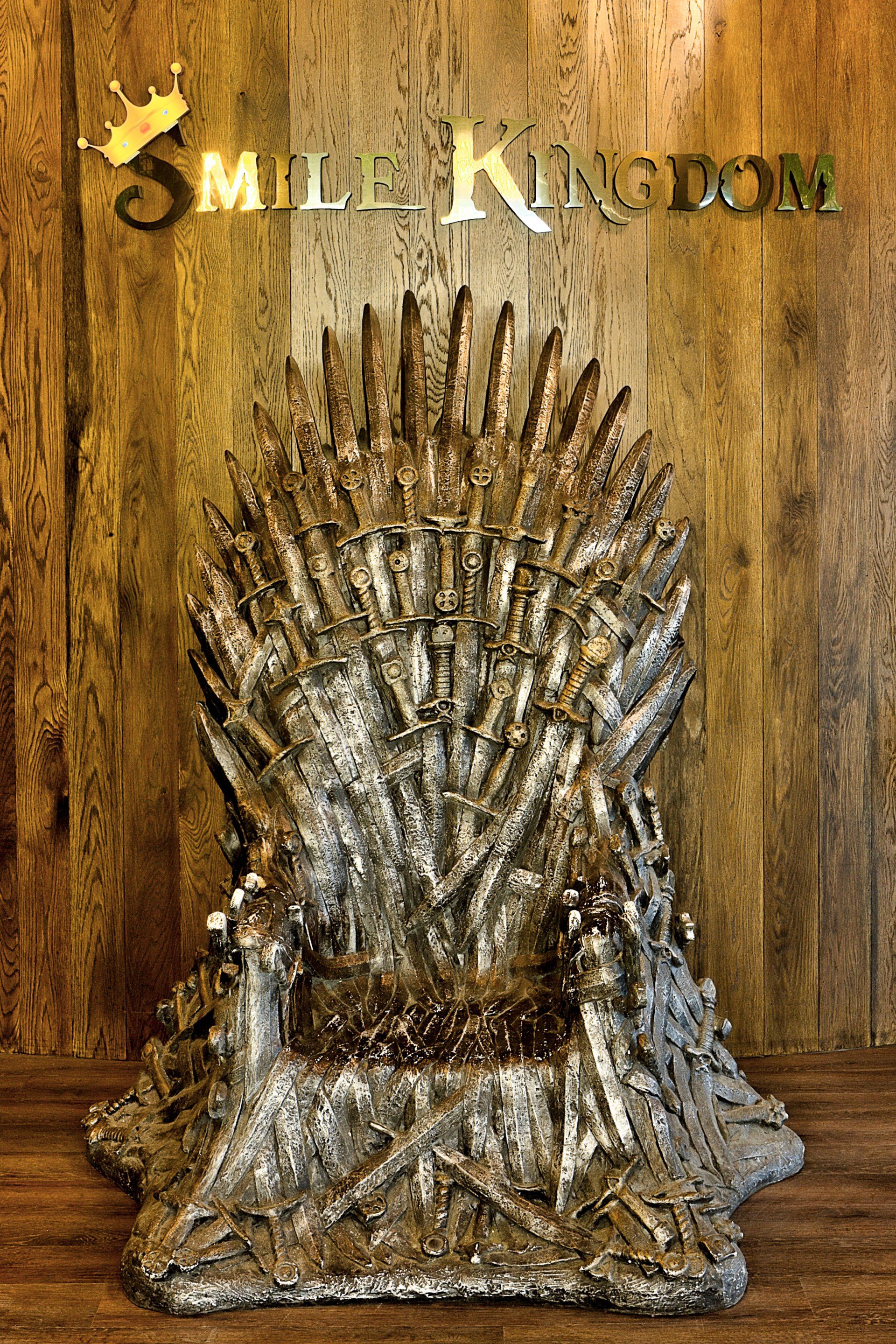 Throne made of silver swords and daggers against a wood wall with “SMIL KAT…” text above