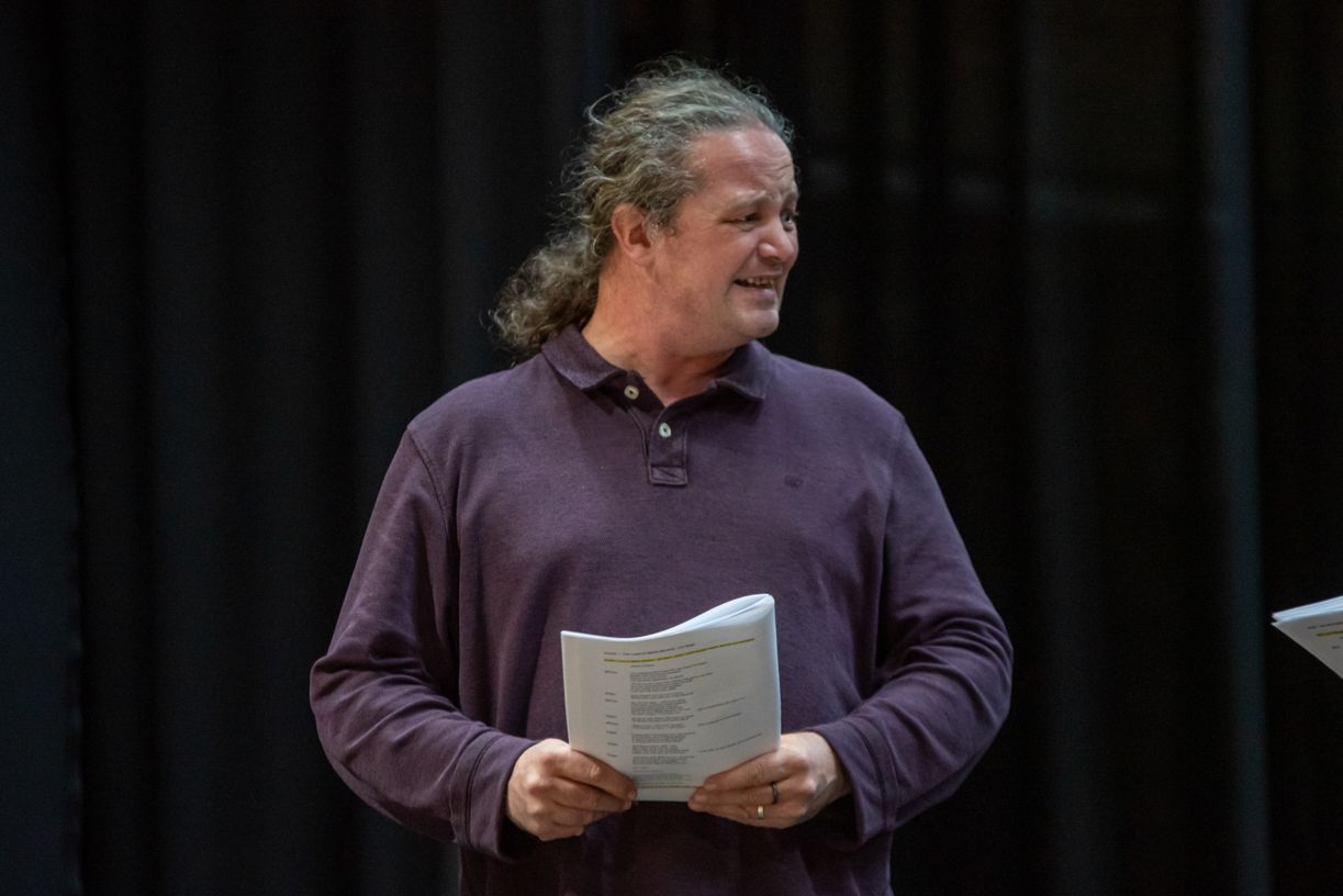 A man in a purple shirt is holding a piece of paper in front of a black curtain.