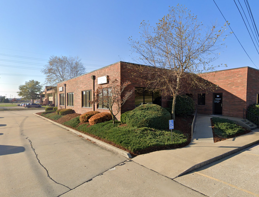 Brick office building beside a sidewalk and drive, with trimmed shrubs and bare trees on a sunny day