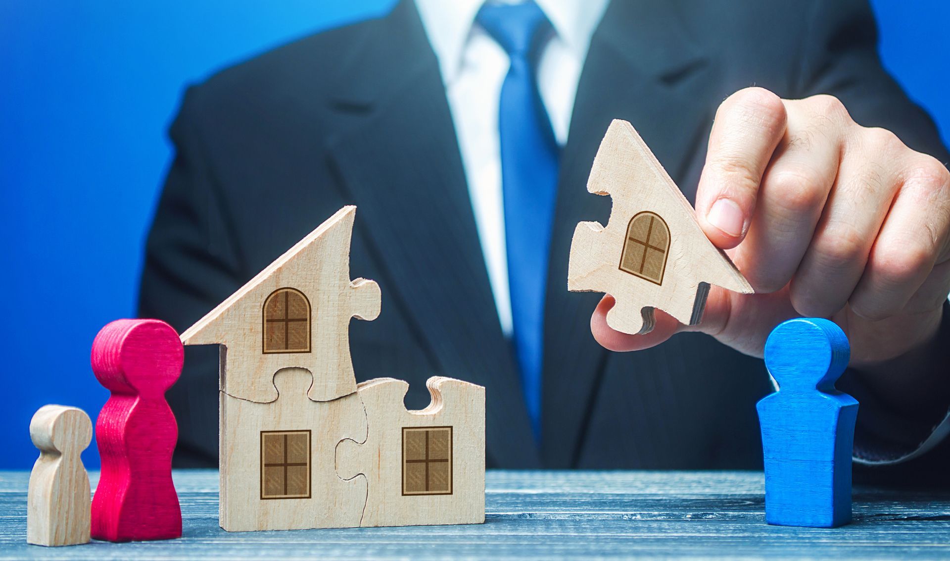 A man in a suit and tie is holding two wooden puzzle pieces in front of wooden houses.