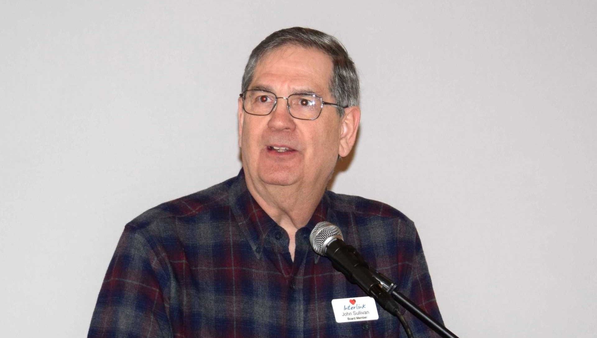 Man speaking at a microphone, wearing glasses and a dark plaid shirt against a plain background