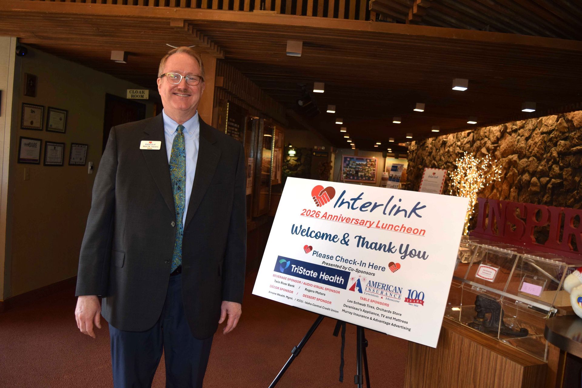 Man in a suit stands beside a thank-you sign at an Interlink event in a wood-paneled room