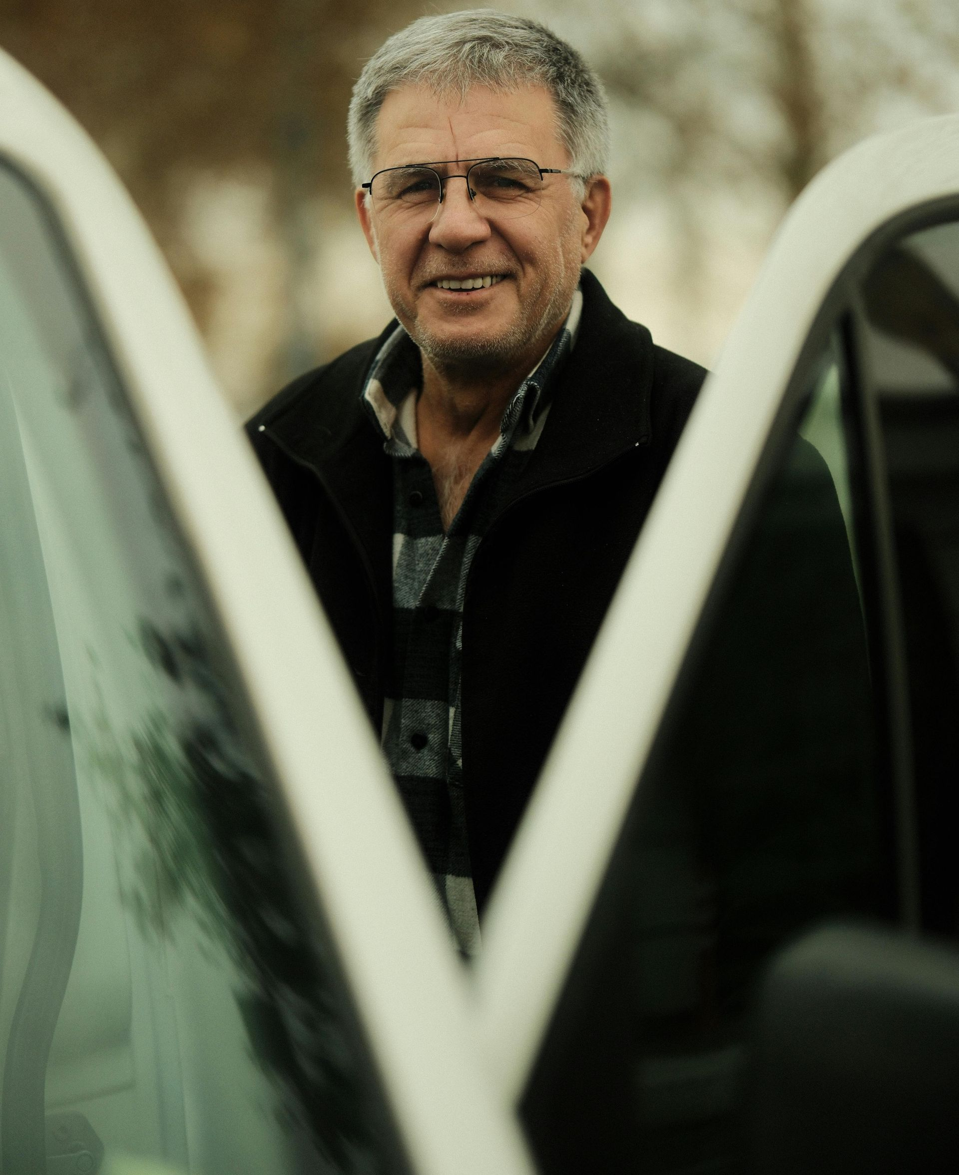 Man standing between two car doors, wearing a dark jacket and glasses, looking at the camera.