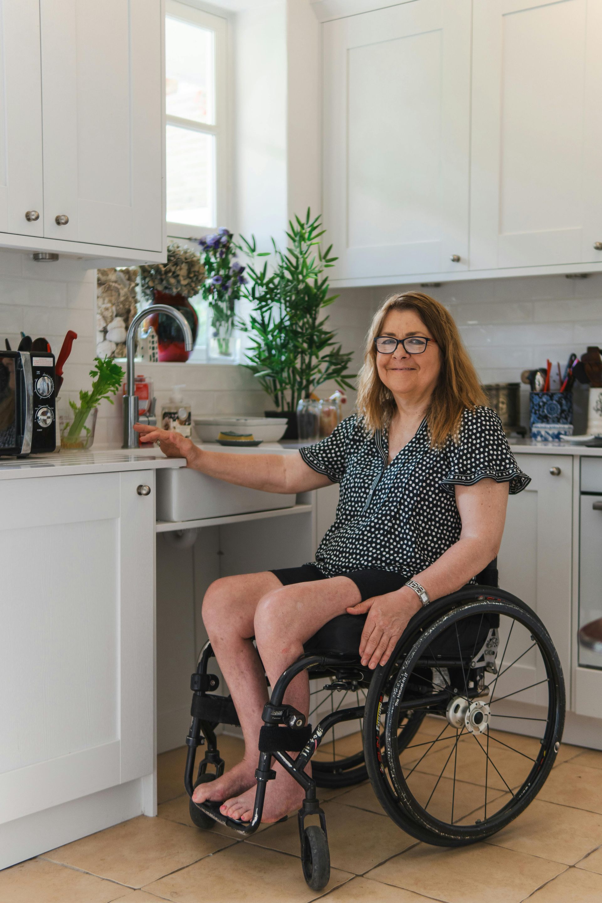 Woman in a wheelchair smiling in a bright kitchen with white cabinets and potted plants