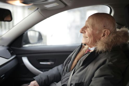 Elderly passenger sitting in a car, wearing a dark coat with a fur collar, looking out the window