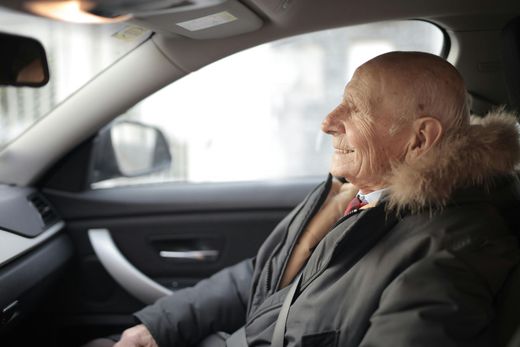 Elderly passenger sitting in a car, wearing a dark coat with a fur collar, looking out the window