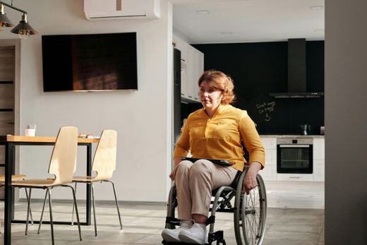 Person in a wheelchair outdoors near a bench and red kiosk, wearing a beige coat and blue jeans.