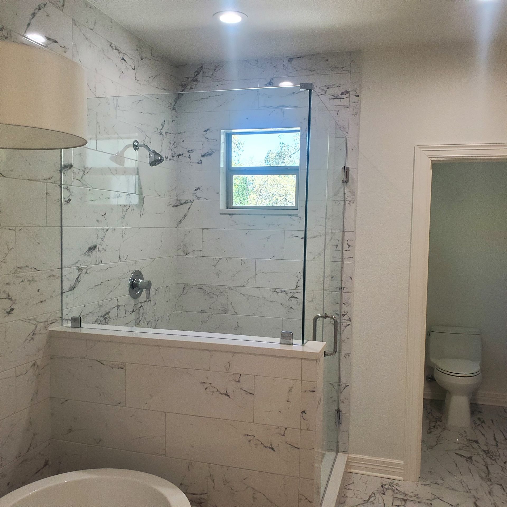 A modern bathroom with a glass shower enclosure and white marble walls. A toilet is visible in the background.