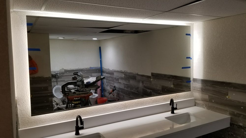 Bathroom under construction with large rectangular mirror, LED lighting, two sinks, and gray-tiled accent wall.