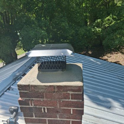 Brick chimney with metal cap on a corrugated roof, surrounded by green trees.