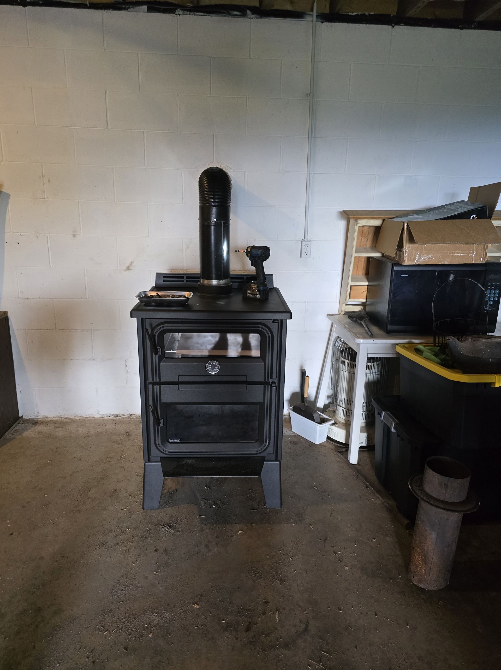 Black wood stove in a bare workshop against a white wall, with tools and shelves to the right.