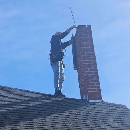 Worker on a shingled roof cleaning a brick chimney against a blue sky.