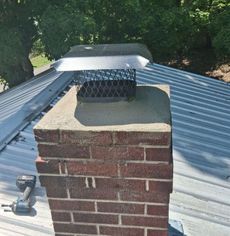 Brick chimney with metal cap on a corrugated metal roof, surrounded by trees.
