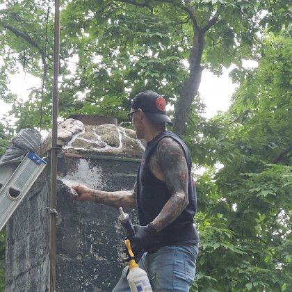 Worker with tattooed arm painting a stone wall outdoors among trees