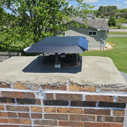 Brick chimney cap with black metal vent on a sunny rooftop, with a house and trees in the background