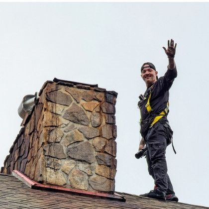Worker on a rooftop beside a stone chimney, waving and wearing a safety harness.