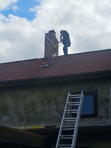 Worker on roof repairing a chimney beside a ladder under a cloudy sky