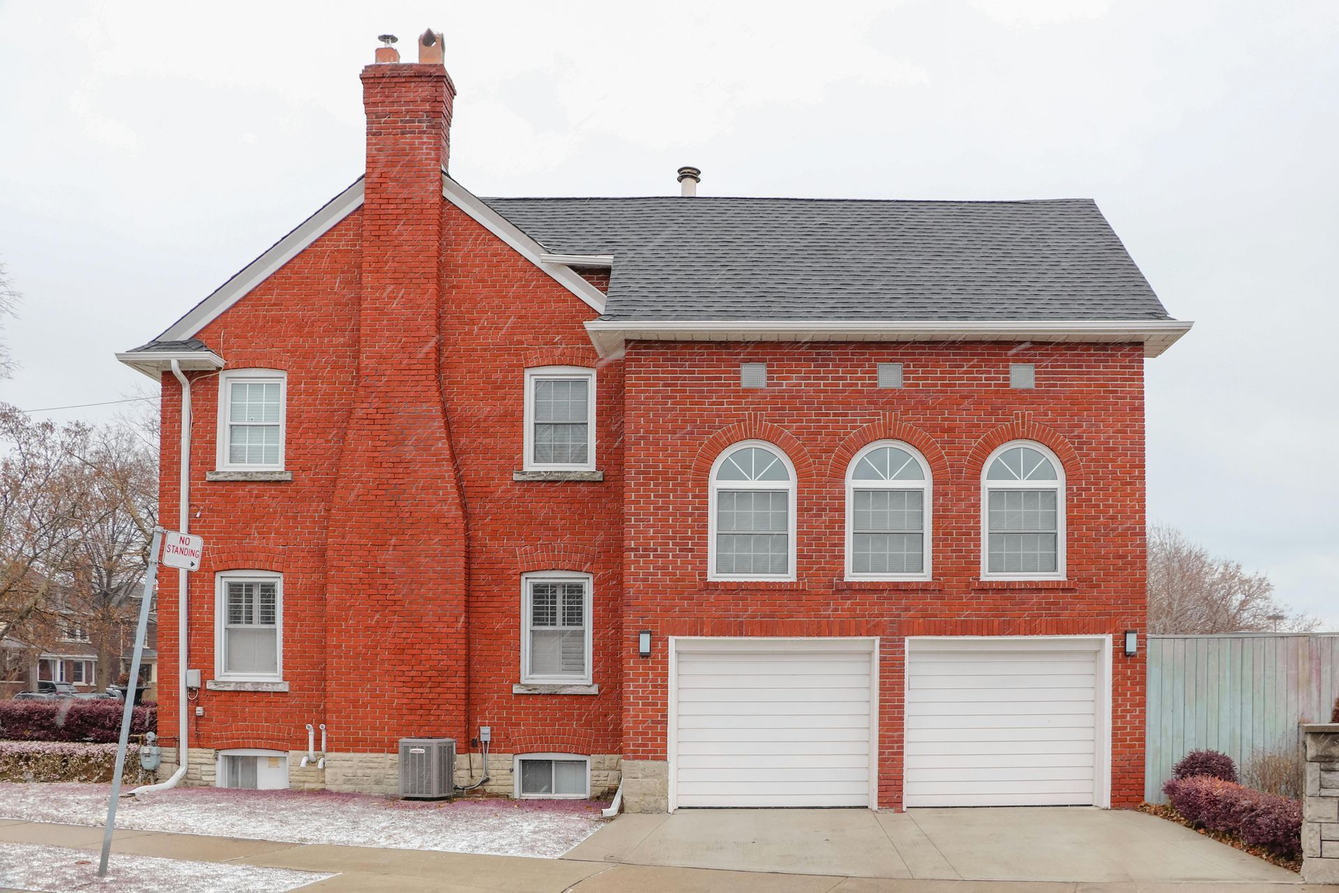 Red brick two-story house with two white garage doors and a tall chimney under a gray roof