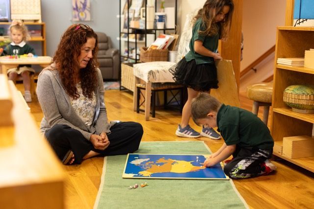 Teacher and child working on a map puzzle on the floor in a classroom, with shelves nearby