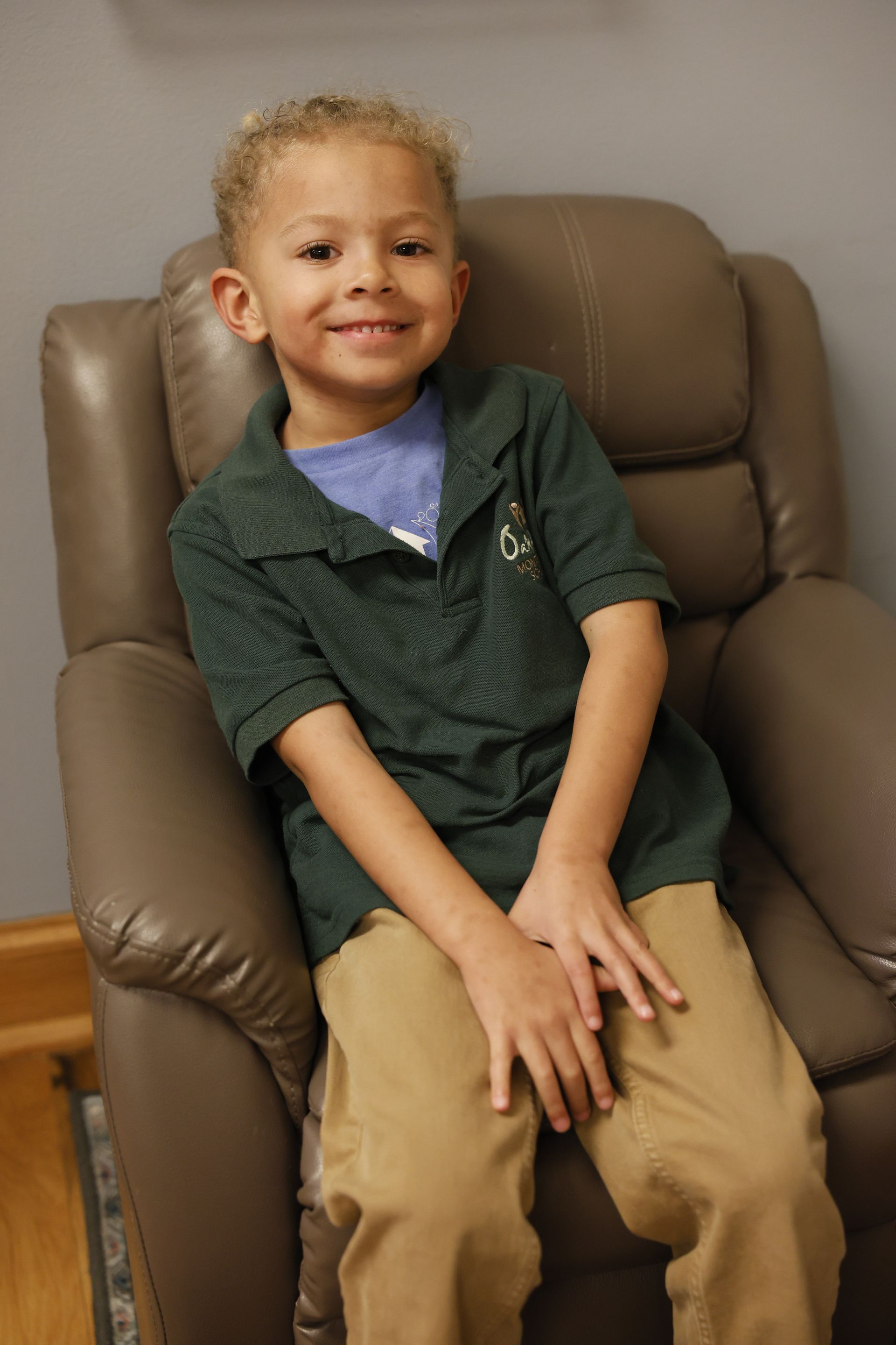 Smiling child sitting in a brown armchair, wearing a green shirt and tan pants.