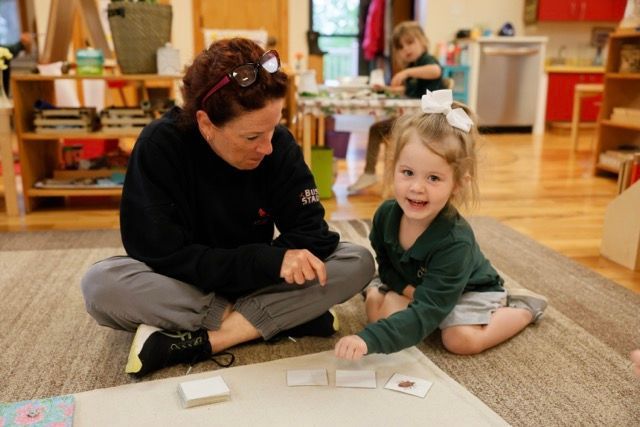 Two people sit on a rug in a classroom, smiling while looking at cards on the floor.
