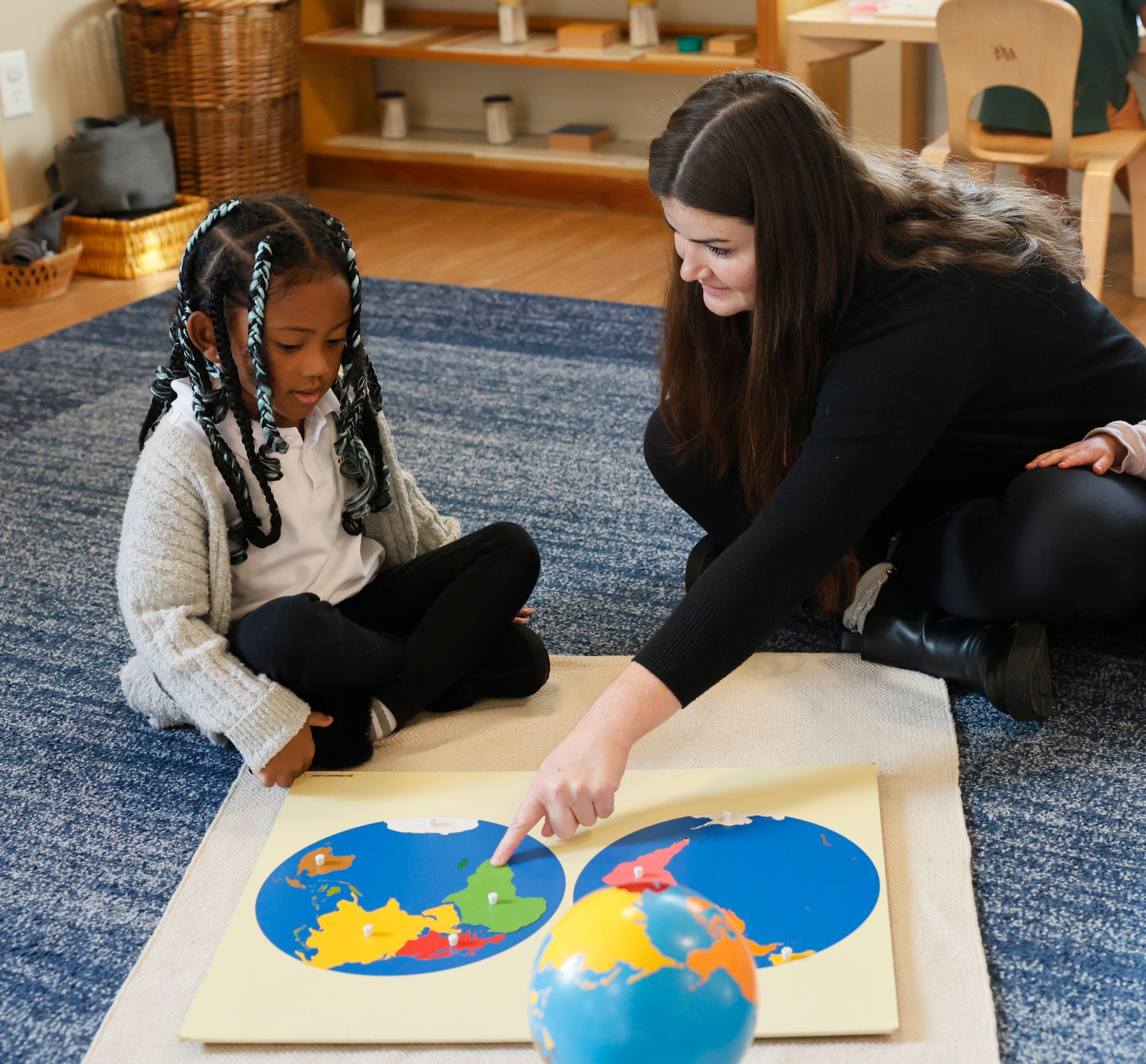 Teacher and child on a rug, pointing at a colorful puzzle map of the world