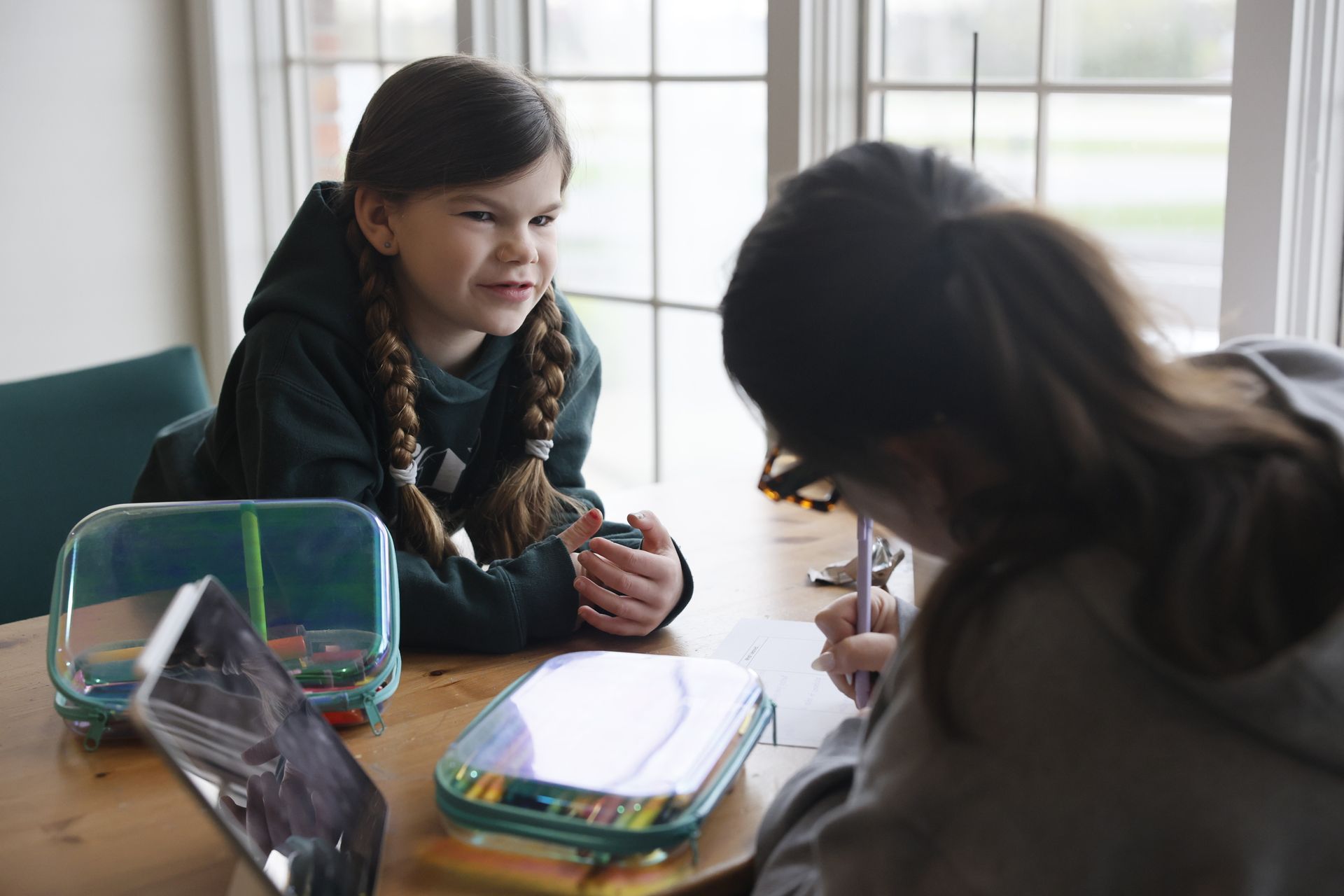 Two students sitting at a table, one smiling while talking in a classroom setting with notebooks and a pencil case