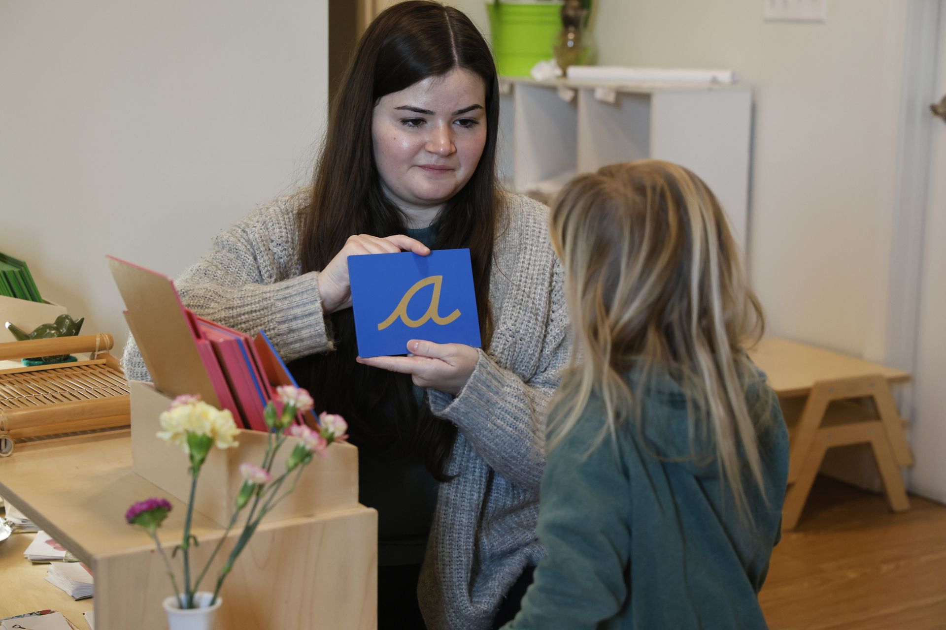 Woman shows a child a blue letter with a yellow A in a classroom setting
