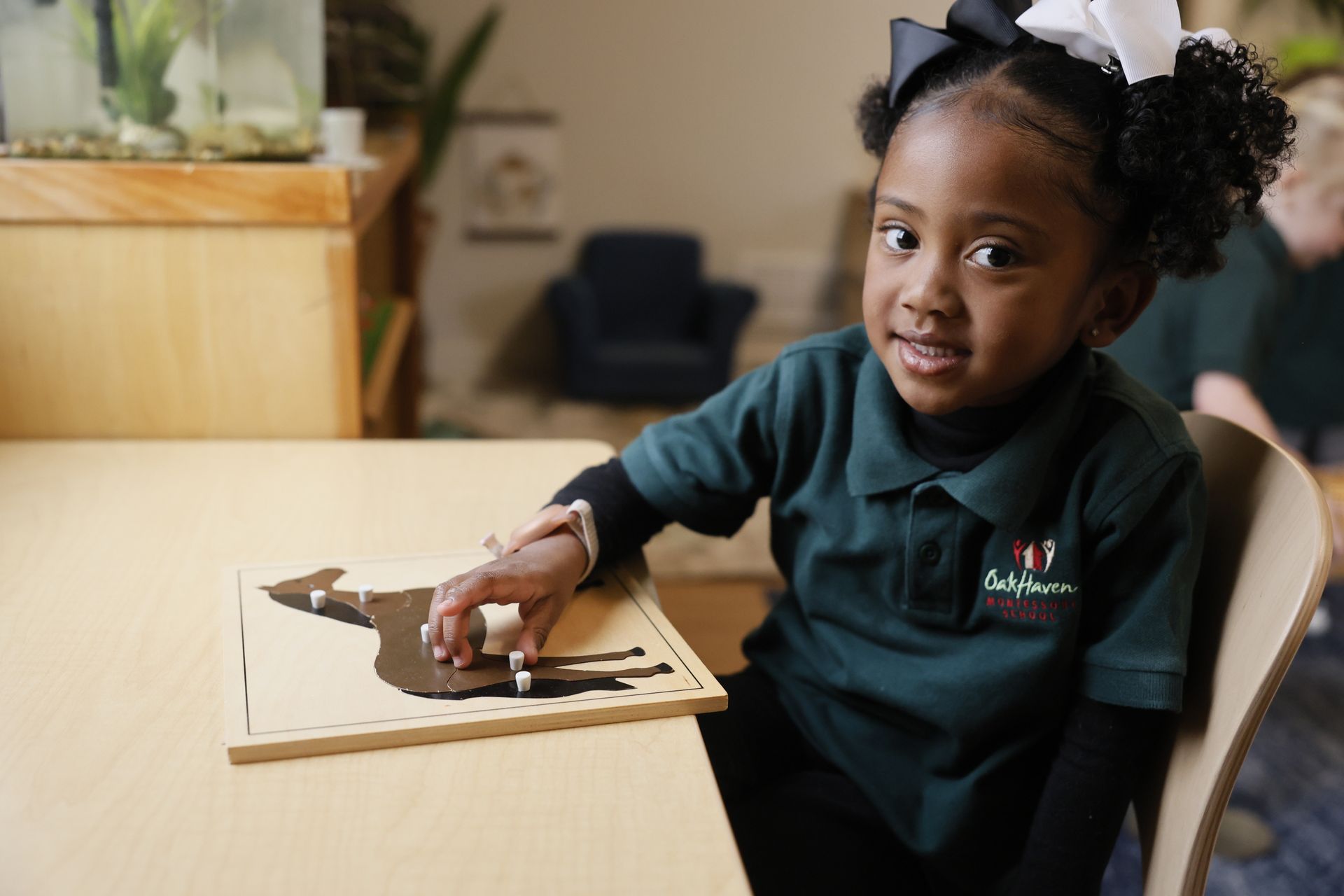 Child in a dark green shirt sits at a table, smiling at the camera.