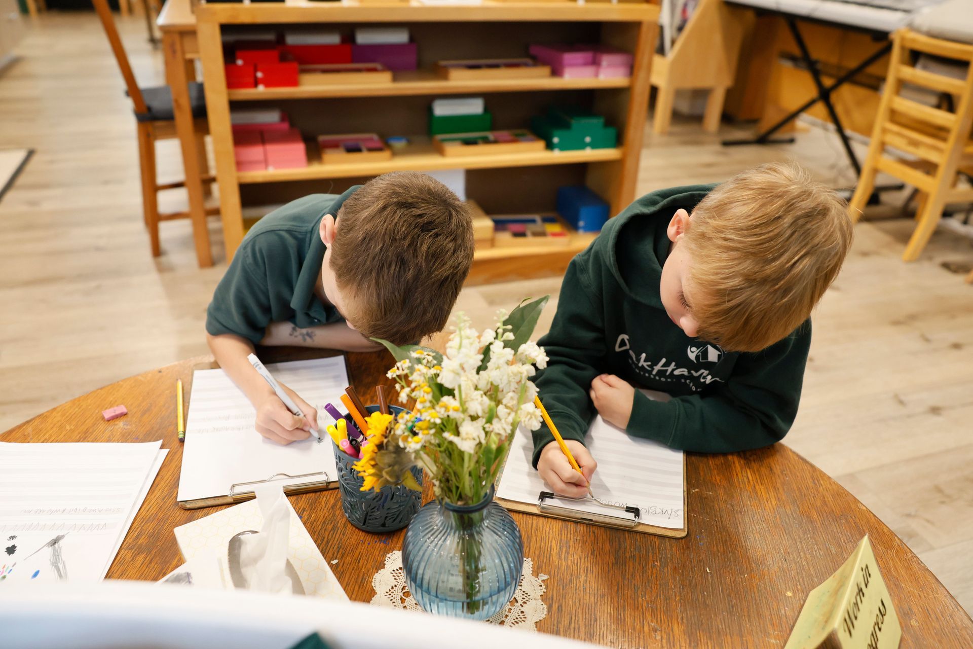 Two children drawing at a table in a classroom, with flowers in a vase in the foreground.
