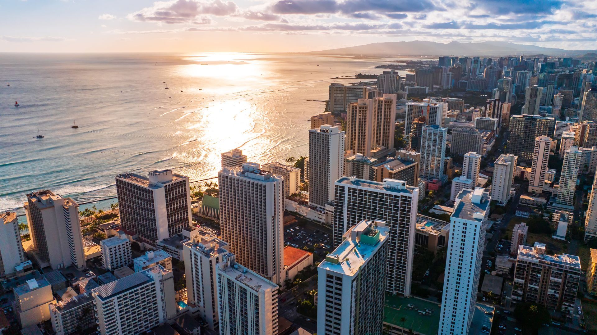 Aerial view of Honolulu, Hawaii, with cityscape and ocean under a bright, setting sun.