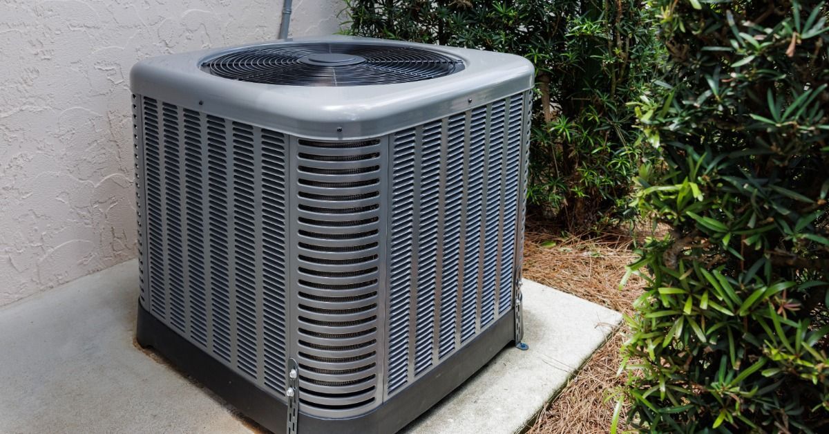 A gray air conditioning unit sitting on a concrete slab beside a stucco wall and green shrubs