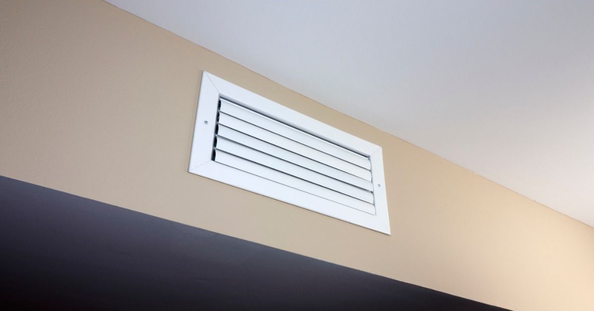 A white rectangular air vent with closed horizontal slats installed high on a beige wall near a white ceiling.