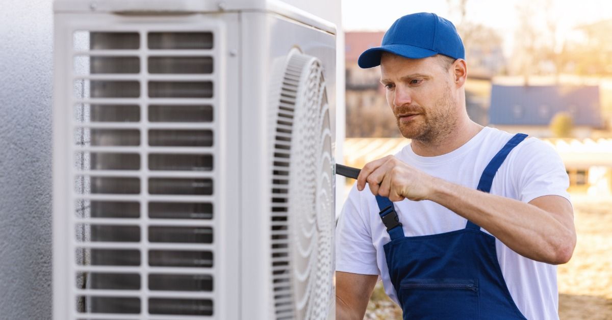 A man in blue overalls and a blue cap holding a tool beside a white outdoor air conditioning unit near a wall.