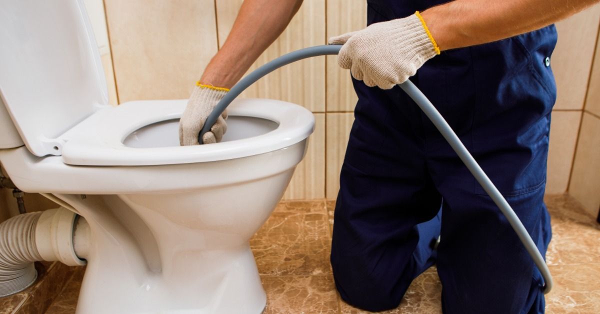 A person in navy work clothes and gloves kneeling beside a white toilet while holding a gray hose in the bowl.
