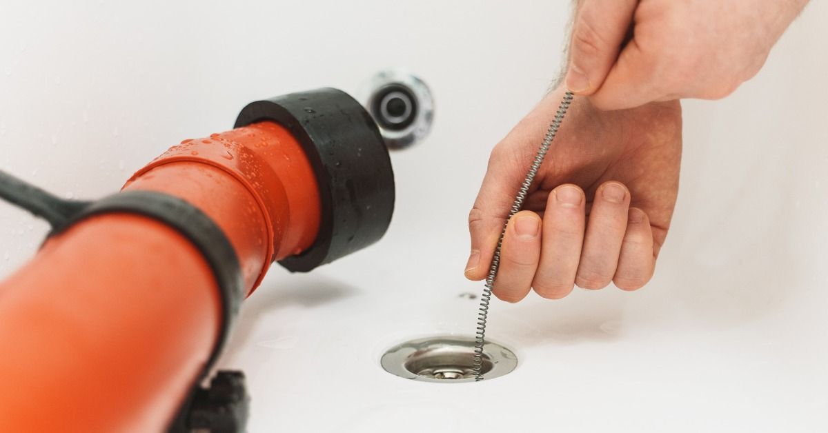 A hand feeding a metal drain snake into a white bathtub drain beside an orange plumbing tool and bla