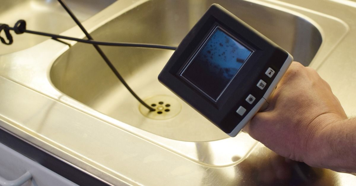 A hand holding a small screen device while a black cable extends into a stainless steel kitchen sink drain.