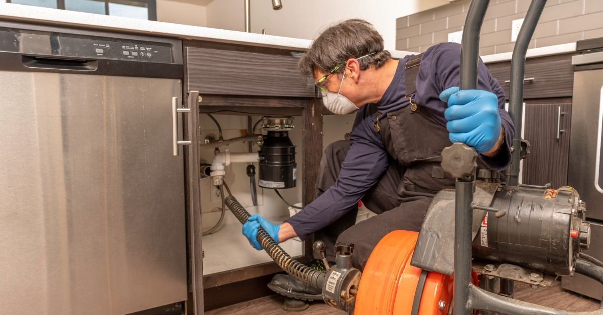 A person wearing a face mask and blue gloves guiding a drain cleaning hose under a kitchen sink beside an orange machine.