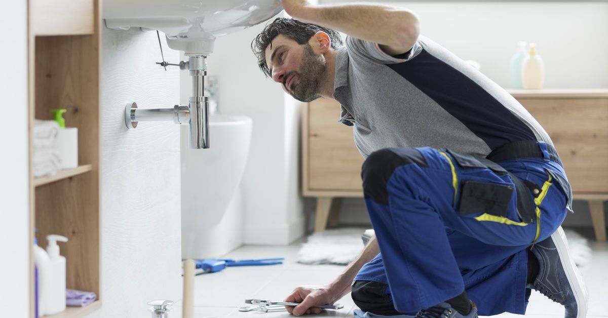 A repairman kneeling under a bathroom sink while checking a pipe. He has some tools sitting next to 