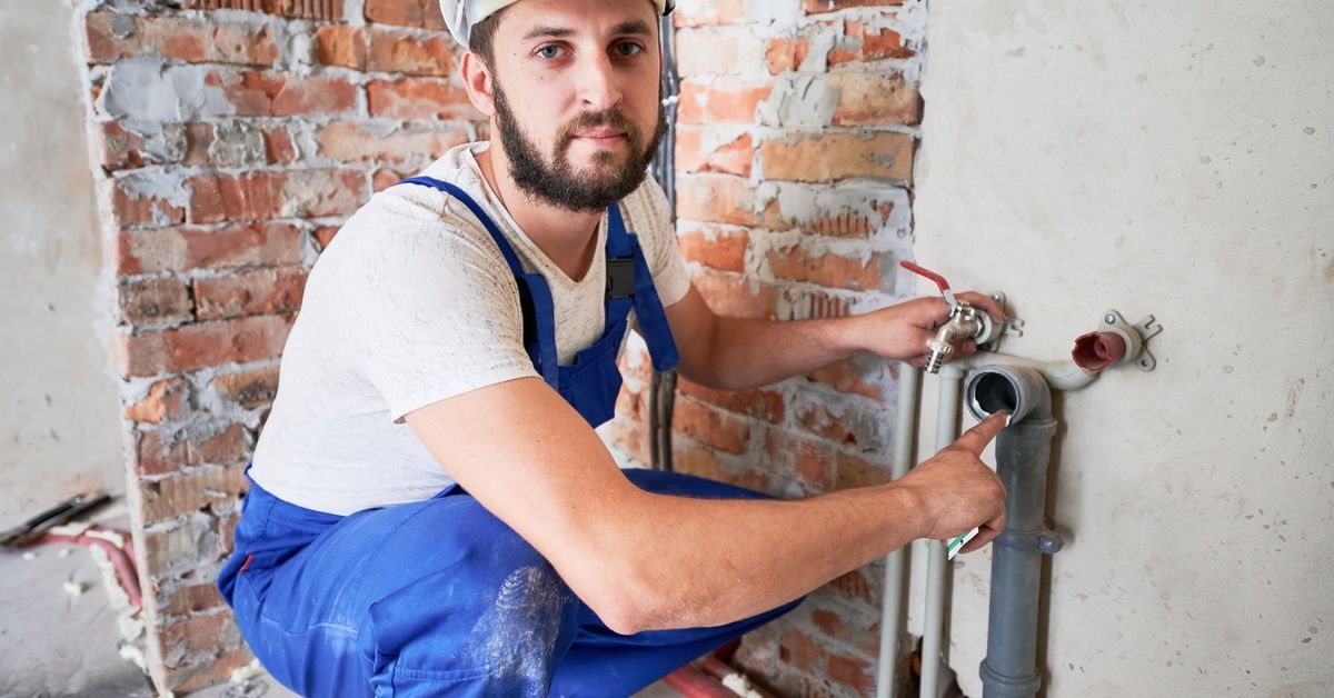 A worker crouched down and pointing at the opening of a pipe. He is wearing a hard hat and blue overalls.