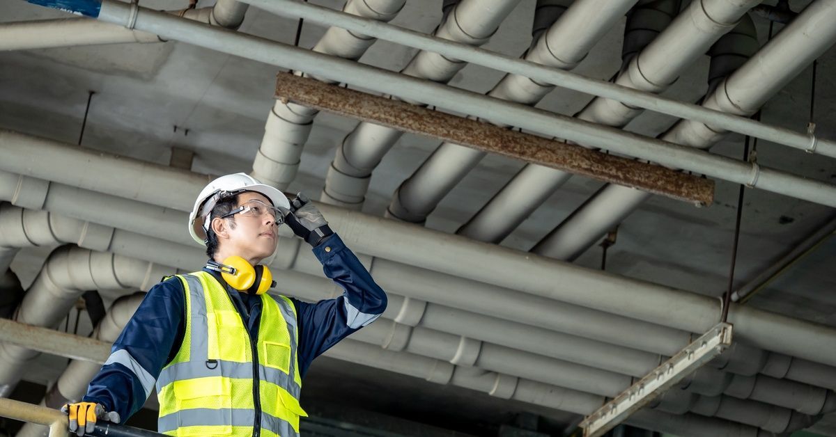 A worker holding his safety helmet while looking up at a large piping system. He is holding onto a nearby railing.