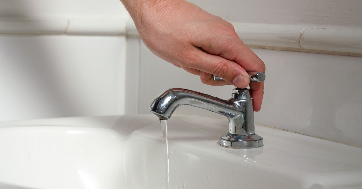A hand turning a valve to turn off a sink faucet. There is a little bit of water still coming out of the nozzle.