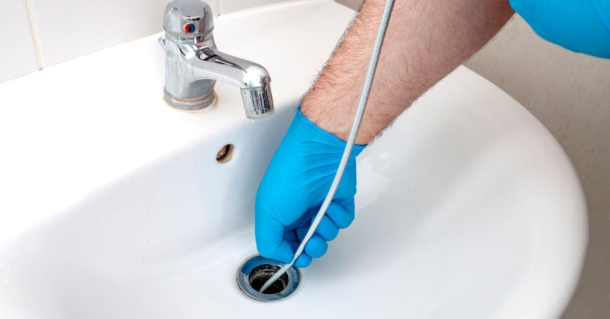A plumber wearing gloves repairs a sink drain using a steel plumber's snake, twisting it through the pipe to clear debris.