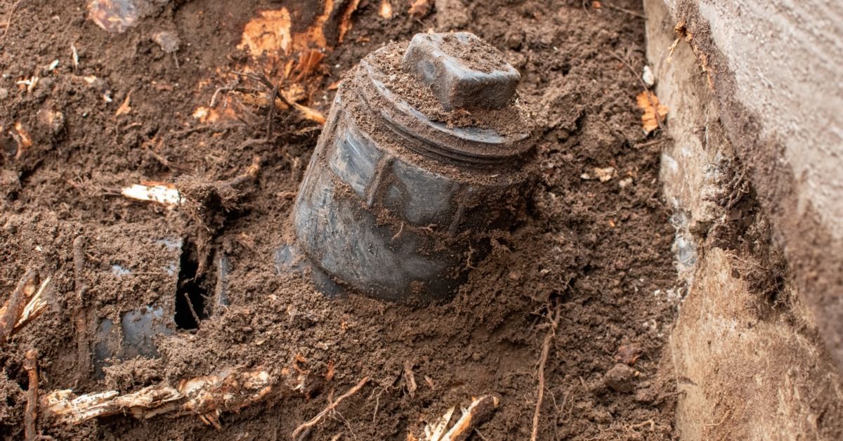 A broken septic pipe lies in excavated soil beside a home foundation, with tree roots wrapped around it.
