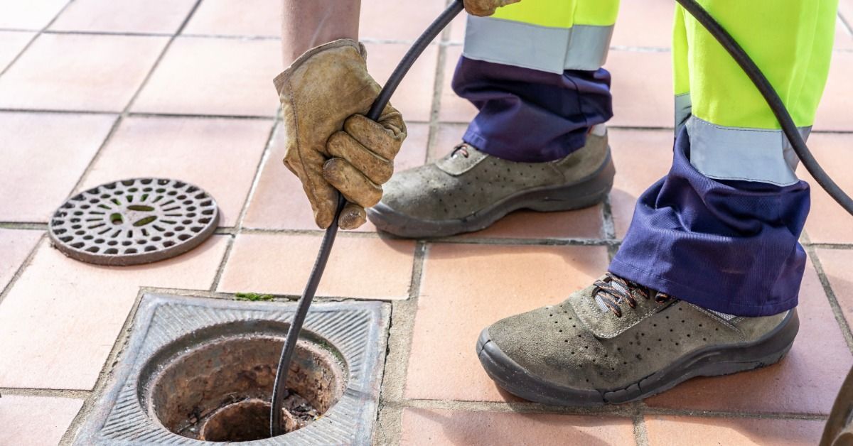 A worker wearing gloves and safety pants guiding a hydro jetting hose into an open drain on tiled ground.
