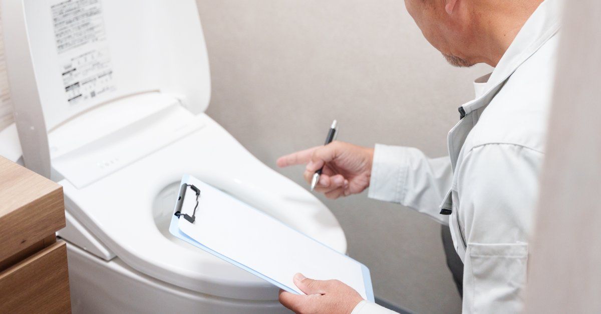 A close-up of a man in a white shirt holding a clipboard and pen as he inspects a toilet with the lid propped up.