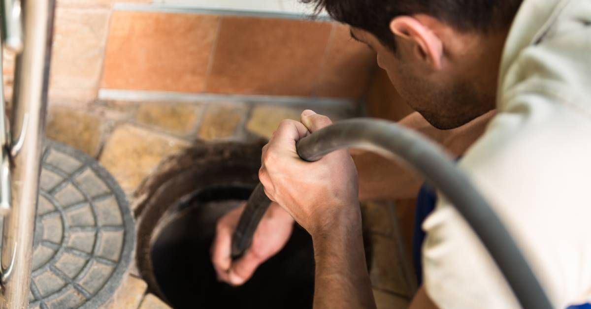 A close-up of a man holding a hose and guiding it into a drainage hole in an orange stone pavement floor.